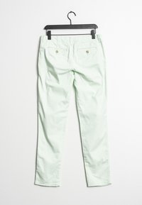 Jasnozielone spodnie o prostym kroju wiszące na czarnym wieszaku, z widocznymi tylnymi kieszeniami z guzikami na białym tle.