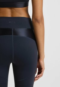 Woman wearing black athletic leggings and crop top, back view, hand relaxed by side, "BOSS" logo on waistband.