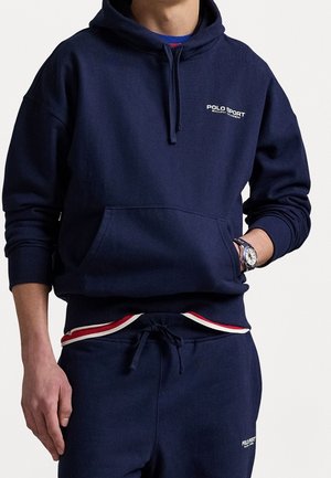 Mężczyzna ubrany w granatową bluzę Polo Sport z kapturem i dopasowane spodnie dresowe z pasem w paski w kolorach czerwonym, białym i niebieskim, zegarek na lewej ręce.