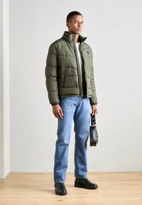 Green padded jacket with a high collar, zip closure, and side pockets. Worn over a white shirt with blue jeans and black shoes.