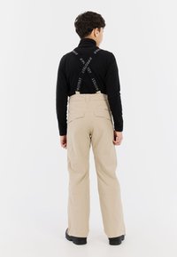 Beige sneeuwbroeken met verstelbare zwarte bretels, een recht model en achterzakken, gedragen over een zwart shirt met lange mouwen.