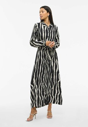 Femme portant une robe maxi à rayures zèbre noires et blanches, à manches longues et avec lien à la taille, posant dans des sandales à talons hauts argentées.