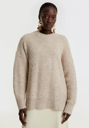Beige gebreide trui met geribbelde textuur, ronde hals en verlaagde schouders. Soepele stof met subtiele kleurvariatie.