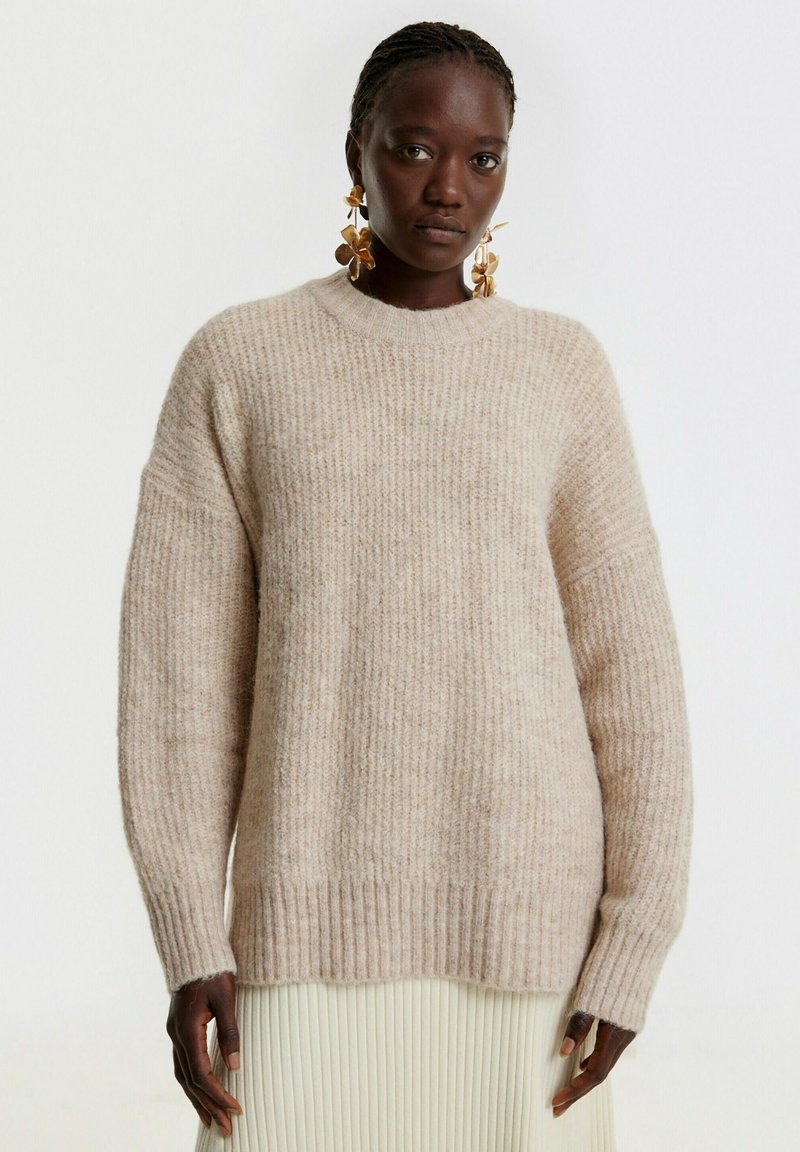 Beige Strickpullover mit geripptem Muster, Rundhalsausschnitt und überschnittenen Schultern. Glattes Material mit subtilen Farbvariationen.