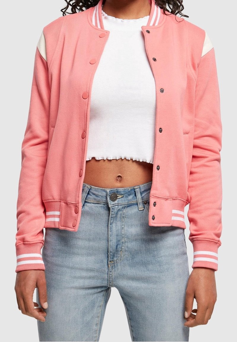 Femme portant une veste varsity rose par-dessus un top court blanc et un jean taille haute bleu clair, debout les mains détendues le long du corps.