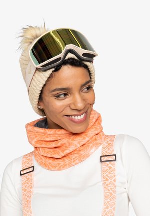 Beige gebreide muts met een pompon, oranje gemusterde nekwarmer en skibril met spiegelglazen, gedragen over een witte top.