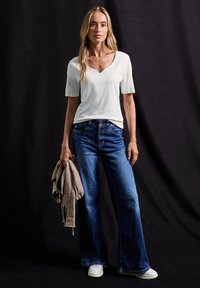Bílý v-neck tričko, modré široké džíny, modelka držící béžovou mikinu, bílé tenisky. Hladká tkanina, volný střih, denim středního praní.