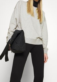 Donna che indossa un felpa grigia chiara e pantaloni neri a coste tiene una borsa nera trapuntata con una larga tracolla contro uno sfondo bianco.