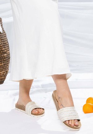 Tan platform sandals with textured white straps, open toe, and a rubber sole. Worn with a flowing white dress.