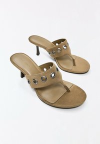 Sandales à talons hauts en daim beige avec boucle pour l’orteil et large bride ornée de détails œillets argentés sur fond blanc.