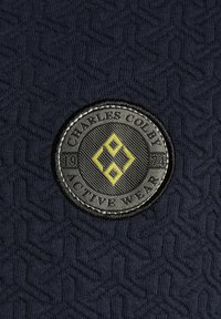 Patch rond noir et jaune du logo Charles Colby Active Wear sur un tissu texturé foncé avec un fond à motif géométrique.