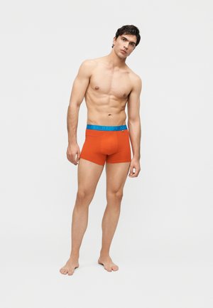 Giovane uomo con capelli scuri in piedi a piedi nudi, indossa boxer arancioni con vita blu con la scritta "HAPPY" su uno sfondo chiaro e uniforme.