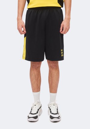 Férfi fekete és sárga sport sortban, fehér zokniban, fekete-fehér sportcipőben, kezeit oldalt tartva áll.