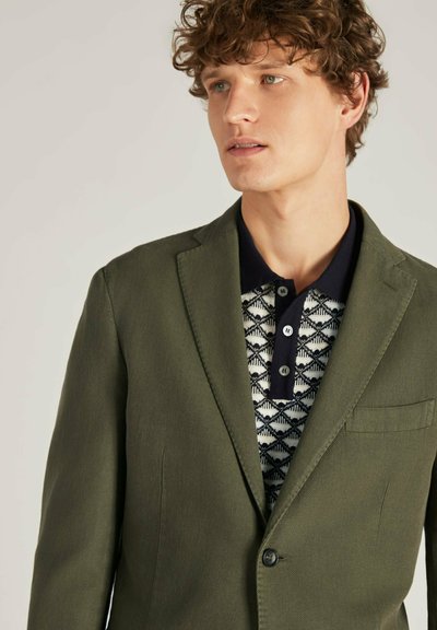Blazer vert olive avec une finition texturée, fermeture à un bouton, et une poche poitrine, superposé à un polo bleu marine à motifs.