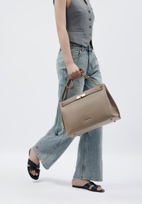 Beige Lederhandtasche mit strukturiertem Design, glänzenden Beschlägen, seitlichem Griff und Metallakzenten, gehalten von einer Person in lässiger Kleidung.