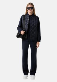 Navy gesteppte Weste mit Druckknöpfen, kombiniert mit einer passenden Bluse und Hose, ergänzt durch eine Handtasche mit schwarzen Fellakzenten und übergroßen Sonnenbrillen.