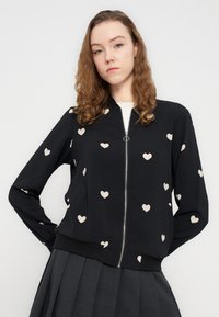 Μαύρο μπουφάν μπότερ με μπεζ σχέδια καρδιάς, κλείσιμο με φερμουάρ και ριμπ μανσέτες και τελείωμα. Φορεμένο πάνω από μια πλισέ γκρι φούστα. Μαλακό ύφασμα.