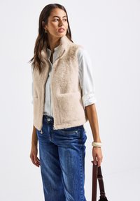 Gilet in finta pelliccia beige chiaro, con zip frontale, due tasche anteriori e colletto. Abbinato a jeans a gamba larga blu e una camicia bianca.