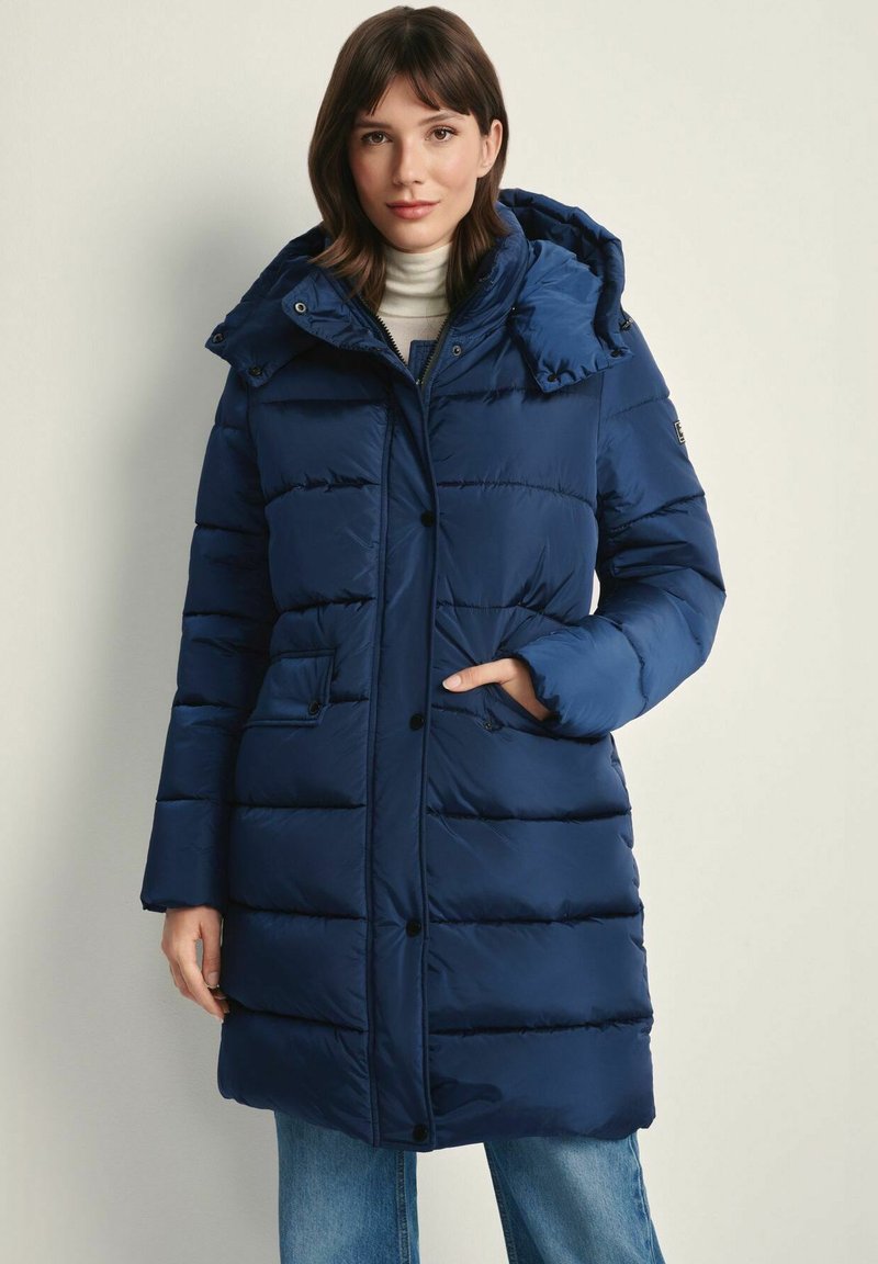 Granatowa kurtka puchowa z odpinanym kapturem, poziomymi pikowaniami, kieszeniami z przodu i zapięciem na guziki, noszona na lekkim swetrze.