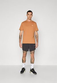 Rjava bombažna majica s kratkimi rokavi, z logotipom na prsih, kombinirana s črnimi kratkimi hlačami in črnimi supergami. Model stoji na enobarvnem ozadju.