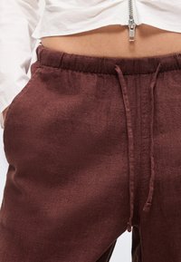 Κοντινό στη μέση που φοράει μπορντό παντελόνι με κορδόνι και ένα λευκό κοντό φερμουάρ που αφήνει ακάλυπτη την περιοχή της κοιλιάς.