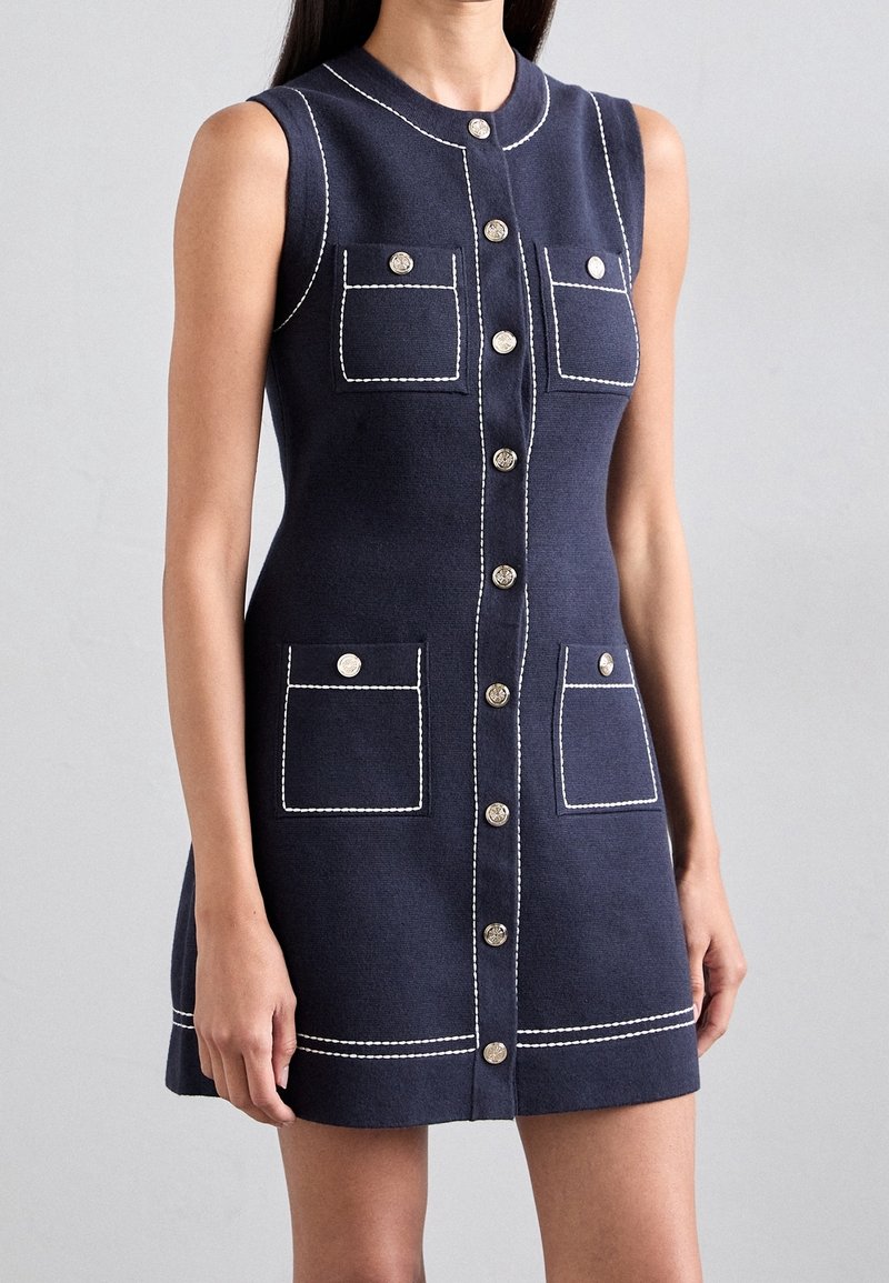 Robe sans manches bleu marine avec boutons sur le devant, couture blanche contrastée et quatre poches à rabat, portée par une personne debout.