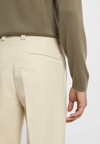 Beige broek met achterzakken, gestructureerde stof en een lichte glans, gecombineerd met een olijfgroen gebreid trui die een deel van de arm onthult.