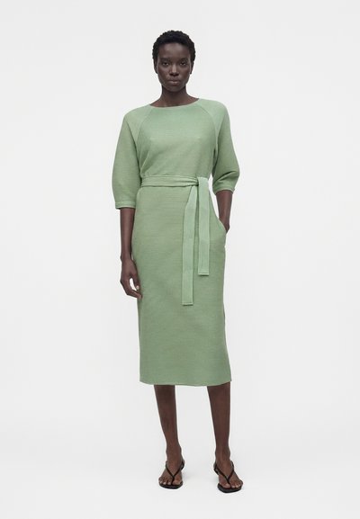 Γυναίκα που φοράει ένα ανοιχτοπράσινο πλεκτό μίντι φόρεμα με μανίκια τριών τετάρτων, ζώνη στη μέση και μαύρα σανδάλια με ανοιχτά δάχτυλα, στέκεται μπροστά σε λευκό φόντο.