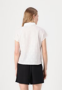 Blusa bianca a maniche corte con motivi floreali ricamati, colletto e vestibilità leggermente morbida, abbinata a pantaloni corti neri.