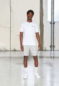 Baltas Champion marškinėlis su logotipu ant krūtinės, derinamas su pilkais šortais, baltais kojinėmis ir baltais sportiniais bateliais; stovintis viduje.