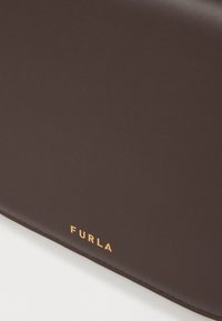 Καφέ δερμάτινη τσάντα με λείο υφής; διαθέτει χρυσό λογότυπο "FURLA" ανάγλυφο στη βάση; ο σχεδιασμός είναι κομψός και μινιμαλιστικός.