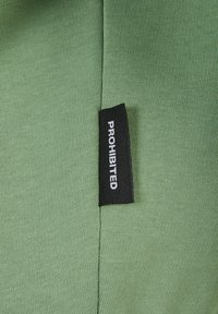 Tissu vert, texturé avec une étiquette noire portant la mention "INTERDIT." Le tissu est doux et présente un design à fines côtes.