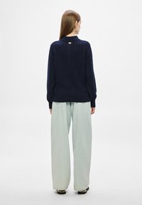 Σκούρο μπλε πουλόβερ με πλέξη κόμπος, μακριά μανίκια, ψηλός γιακάς και χαμηλοί ώμοι· συνδυασμένο με ανοιχτό μπλε παντελόνι φαρδύ.