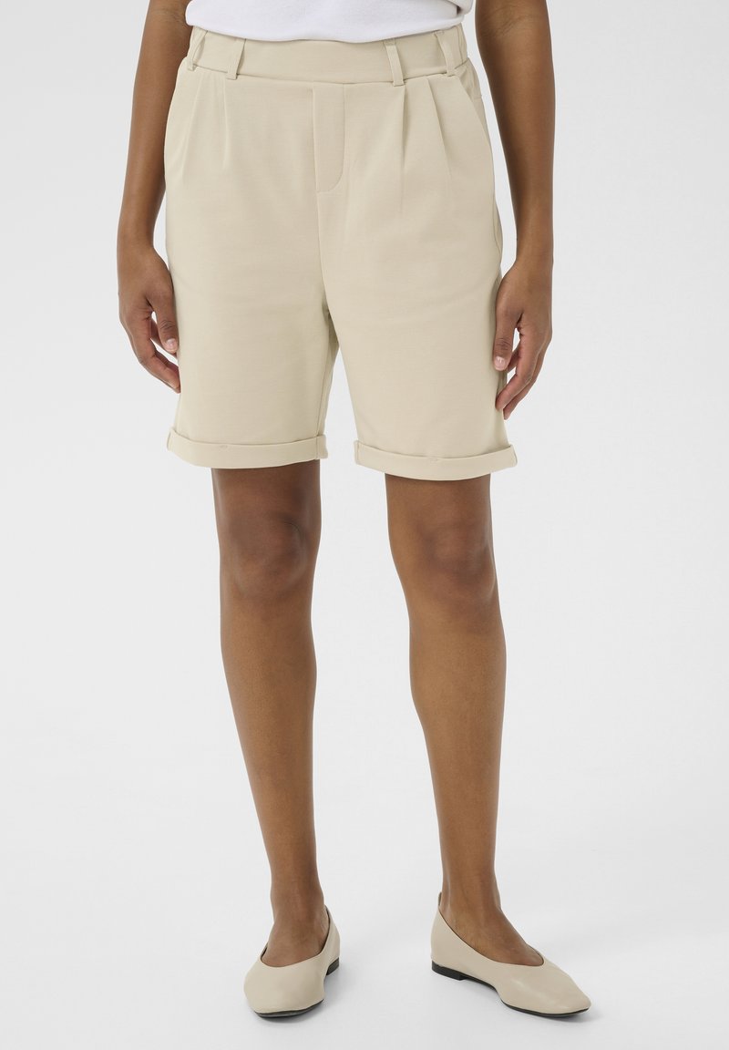 Beige shorts van gladde stof, met voorvouwen en opgerolde zoom. Gecombineerd met beige platte schoenen. Minimalistisch ontwerp.