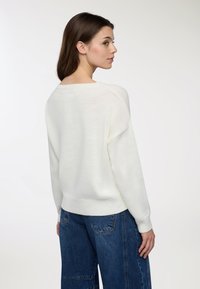 Maglione bianco lavorato a maglia con una vestibilità rilassata, scollatura rotonda e spalle basse. Indossato con jeans blu a gamba larga, che mostrano una morbida trama.