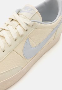 Svetlo krémové kožené tenisky s textúrovaným povrchom, svetlomodrým logom swoosh, plochými šnúrkami a subtílnou béžovou gumovou podrážkou.