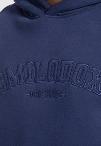 Felpa blu navy con ricamo in rilievo della scritta "SMILODOX" e "ORIGINALS" sul davanti. Tessuto morbido con un design classico.