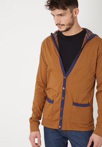 Cardigan marrón con escote en V, ribete azul marino, dos bolsillos frontales y cierre de botones. Fabricado con un tejido de textura suave.