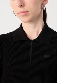 Melns ribots polo krekls ar rāvējslēdzēju un kontrastējošu melnu logo kreisajā pusē. Iespējo klasisku apkakli un teksturētu audumu.