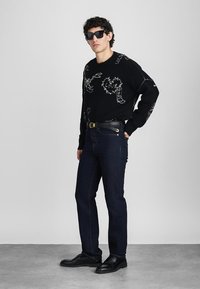 Sweater preto com design gráfico branco, combinado com jeans azul escuro e sapatos pretos. O modelo usa óculos de sol escuros. Fundo minimalista.