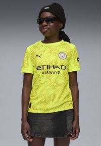 Maglietta da calcio di Manchester City di un vivace giallo con un motivo grafico, logo nero e testo "ETIHAD AIRWAYS". Abbinata a una gonna di jeans.
