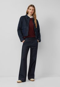 Veste en denim avec fermeture à boutons et poches à rabat, associée à un pull bordeaux et un jean ample foncé, avec des coutures en contraste.