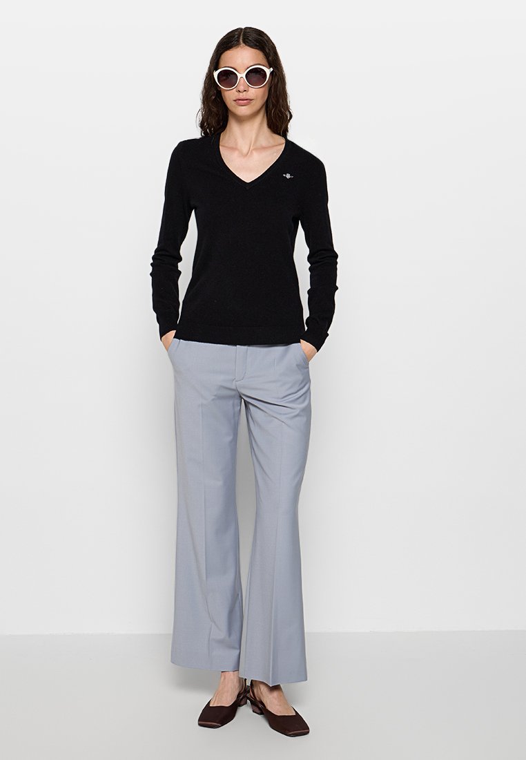 Schwarzer V-Ausschnitt-Pullover, hellgraue weite Hose und dunkelbraune Schuhe. Das Model trägt oversized weiße Sonnenbrillen und hat lange braune Haare.