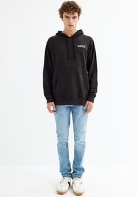 Sudadera con capucha negra con el texto "INSIDE" en el pecho, combinada con jeans de color azul claro y zapatillas blancas con acentos en negro y beige.