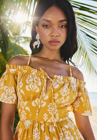 Vestido amarillo con escote off-shoulder y patrones florales blancos, mangas con volantes y un detalle de lazo en el frente. La modelo lleva pendientes florales blancos.