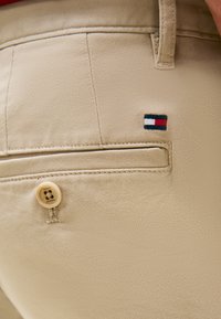 Μπεζ βαμβακερό παντελόνι με πίσω τσέπη που κλείνει με κουμπί και μικρό, κεντημένο λογότυπο σε κόκκινο, άσπρο και μπλε στο πλάι.