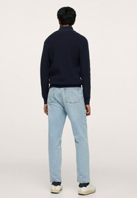 Person in einem dunkelblauen gerippten Pullover, hellblauen Jeans und weißen Sneakers, der mit dem Rücken zu einem schlichten Hintergrund steht.