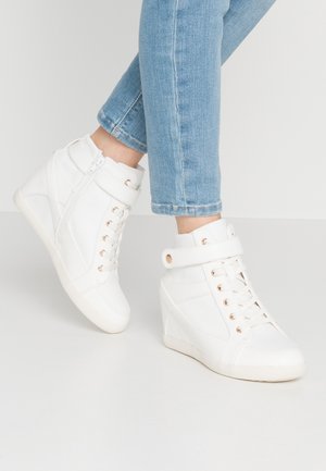 Baskets blanches montantes compensées avec lacets et bride à la cheville, portées avec un jean bleu clair sur fond blanc uni.