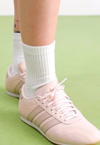 Világos rózsaszín sportcipők fehér fűzővel, a zokni texturált részletekkel a lábujj részen, és három sötét csíkkal az oldalukon, ribbent fehér zoknikkal párosítva.