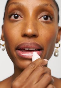 Läppvårdsprodukt hålls nära läpparna, vit tub med rundad spets. Hudtonen är varm, läpparna är lätt tonade i rosa, bär guldörhängen.